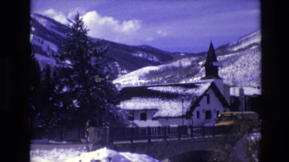 1974 Vail, Colorado 8mm frame with a white church-like building near a bridge, snowy mountain slopes, evergreens, a small distant person, and signs at the roadside.