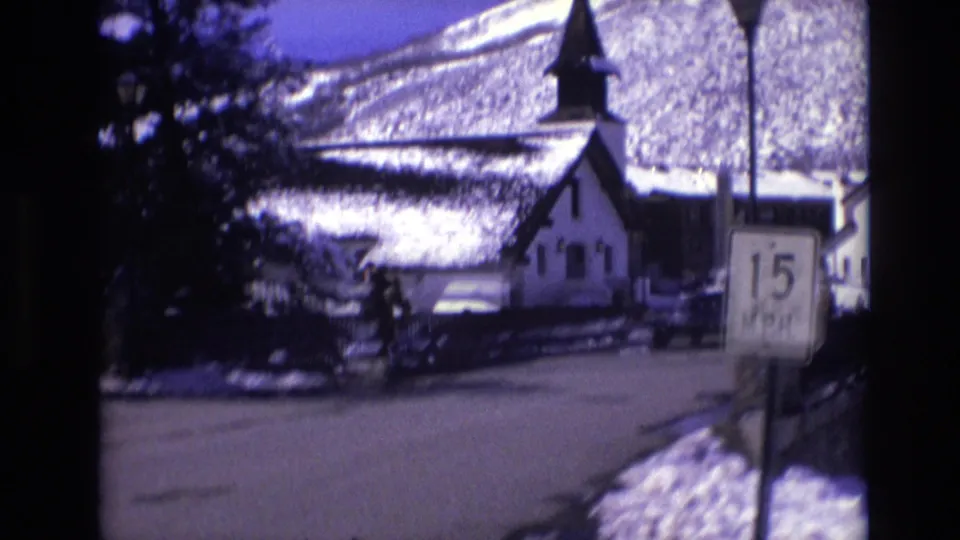 Home-movie frame of a snowy Vail, Colorado road in 1974, with pedestrians near a church-like building, a 15 M.P.H. sign, trees, and snow-covered mountain terrain.
