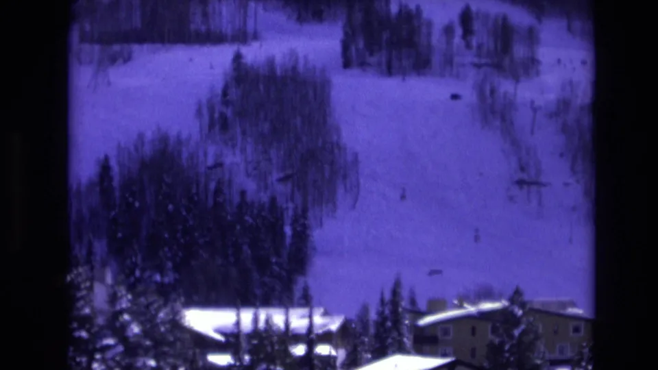 1974 Vail, Colorado view of a snow-covered ski hillside with small distant figures, evergreen trees, and resort buildings along the lower edge of frame.