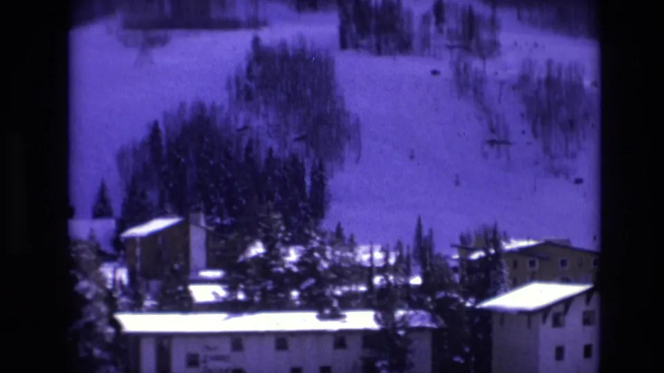 1974 Vail, Colorado frame showing low-rise resort buildings, snow-bright roofs, dense pine trees, and a ski slope rising in the background.