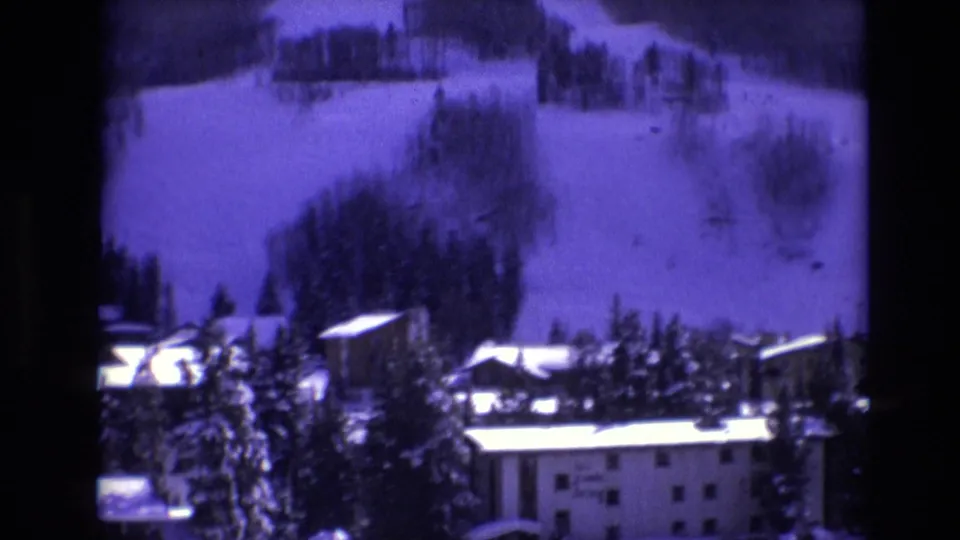 1974 Vail, Colorado home-movie frame with resort rooftops and trees in the foreground, a snowy ski slope behind them, and small distant figures on the hill.