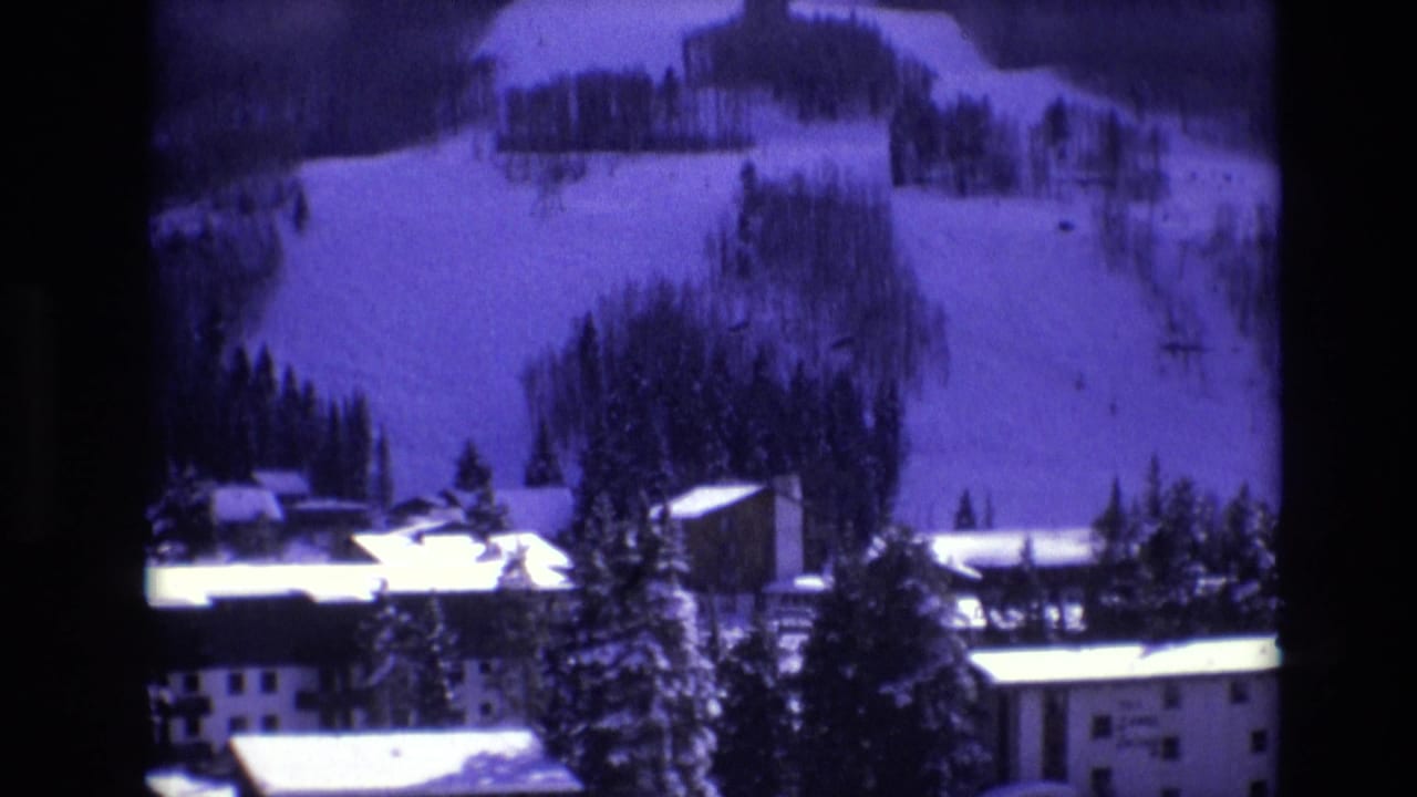 1974 Vail, Colorado home-movie frame showing snow-covered slopes rising behind alpine buildings and dark evergreen trees.