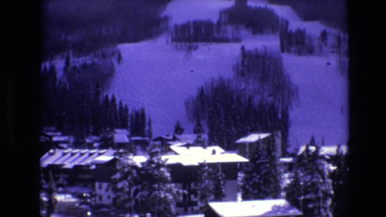 1974 Vail, Colorado frame with snowy resort buildings, evergreens, and ski slopes seen through a soft purple-tinted 8mm image.