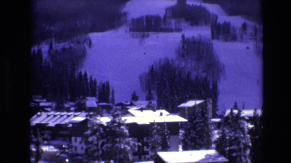 1974 Vail, Colorado frame with snowy resort buildings, evergreens, and ski slopes seen through a soft purple-tinted 8mm image.