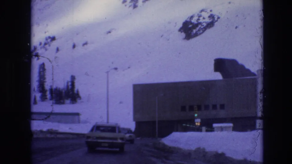 1974 Vail, Colorado road-level frame showing a car approaching a snowy lodge or resort building with mountain snowfields behind it.