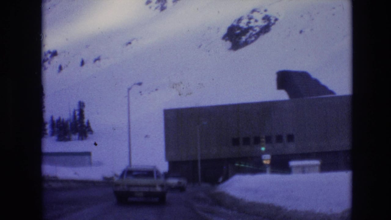 1974 Vail, Colorado 8mm frame showing a car approaching a large concrete underpass with snowbanks, a hillside, and a dark roof form above the structure.
