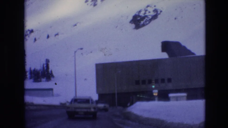 1974 Vail, Colorado 8mm frame showing a car approaching a large concrete underpass with snowbanks, a hillside, and a dark roof form above the structure.