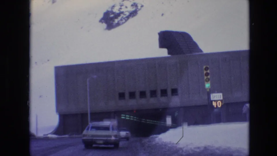 1974 Vail, Colorado 8mm frame showing a station wagon near a concrete underpass, with snowbanks, small upper windows, a green signal, and a speed limit sign.
