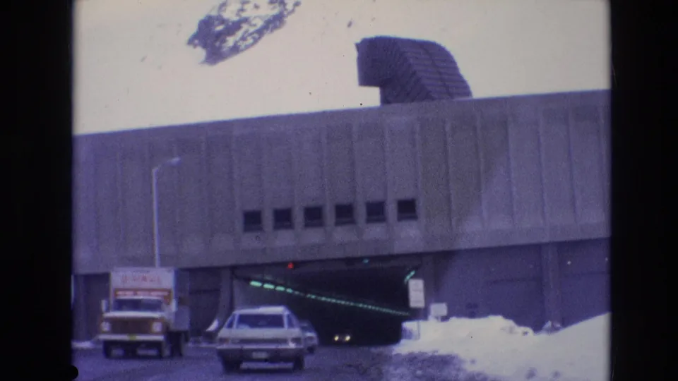 1974 Vail, Colorado frame showing a white truck and a car at the entrance to a large concrete underpass, with snowbanks, illuminated interior lights, and a sculptural roof element.