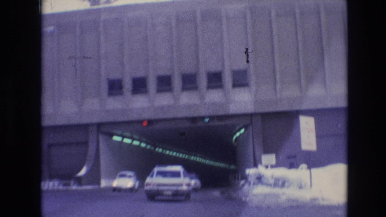 Vehicles move toward a concrete tunnel in Vail, Colorado, with a white station wagon centered near the entrance and snowbanks visible at the edge of the road.