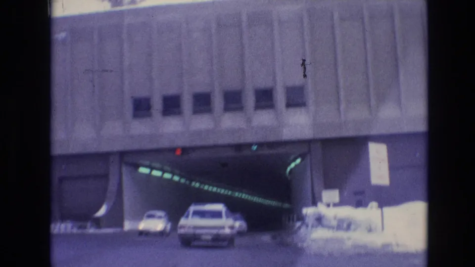 Vehicles move toward a concrete tunnel in Vail, Colorado, with a white station wagon centered near the entrance and snowbanks visible at the edge of the road.