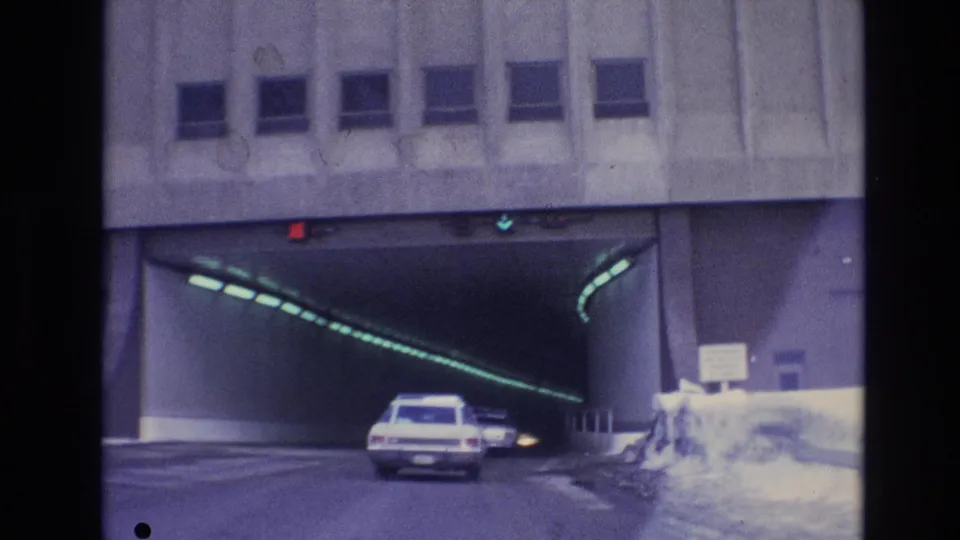 A white 1970s-era car enters the tunnel on a two-lane road, with green lighting curving along the tunnel wall and snow visible outside the entrance.