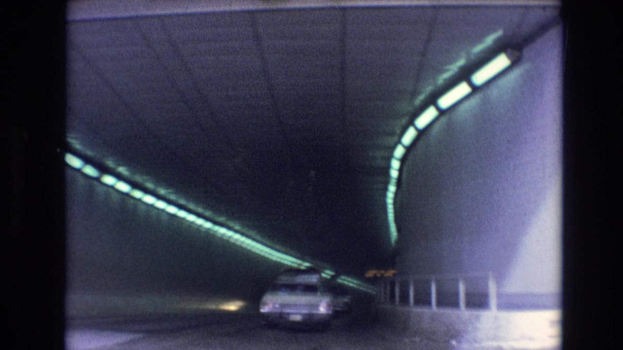 1974 Vail, Colorado home-movie frame showing cars driving through a dark highway tunnel lit by green overhead fixtures.