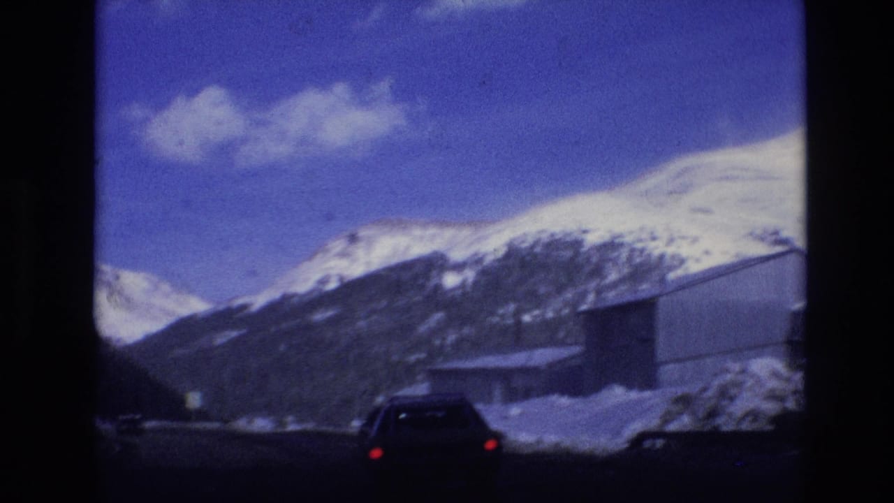 Car on a snowy mountain road with peaks and roadside buildings ahead.