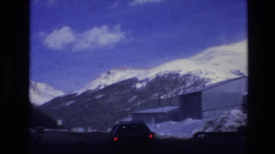 Passenger-view 8mm frame from Vail, Colorado in 1974 showing a car driving along a snow-lined road beneath distant mountain peaks.