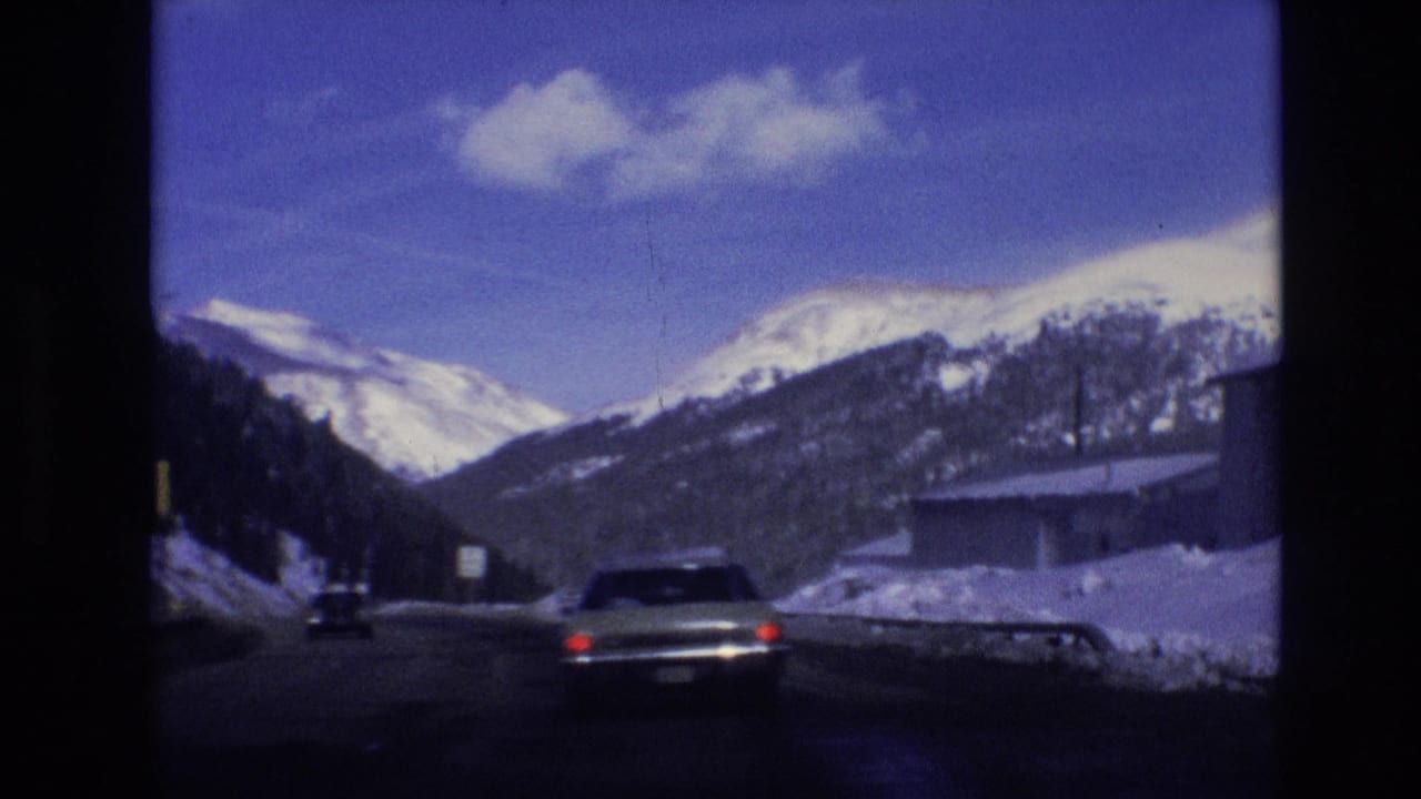 1974 Vail, Colorado road scene with cars moving through a snowy valley, low buildings at the roadside, and snow-covered peaks beyond.