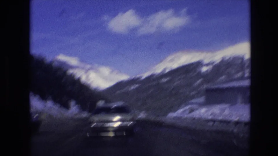 Soft 8mm frame from a moving vehicle in Vail, Colorado showing an oncoming car on a snow-bordered road with mountain slopes behind it.