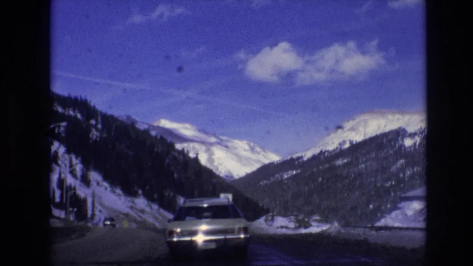 1974 Vail, Colorado home-movie frame showing an approaching car on a winter mountain road, with snowbanks, evergreen slopes, and bright peaks in the distance.