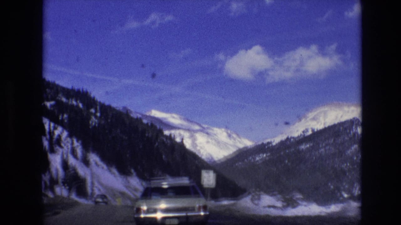 1974 8mm road-trip frame from the supplied Vail, Colorado clip showing a snowy mountain highway, a car ahead, blue sky, and dark film borders.