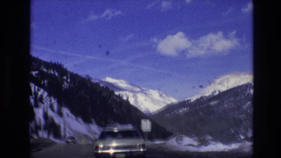 1974 8mm road-trip frame from the supplied Vail, Colorado clip showing a snowy mountain highway, a car ahead, blue sky, and dark film borders.