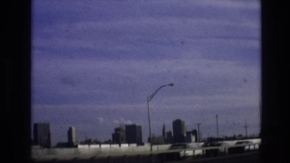 Frame from the supplied 1974 Vail, Colorado clip showing a distant skyline beyond a highway barrier, with streetlights and dark Super 8-style frame edges.