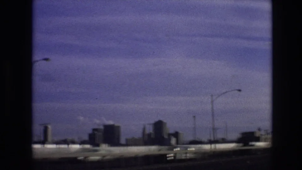1974 8mm moving-car view from the supplied Colorado clip with a skyline in the distance, a highway barrier, streetlights, and motion-blurred traffic.