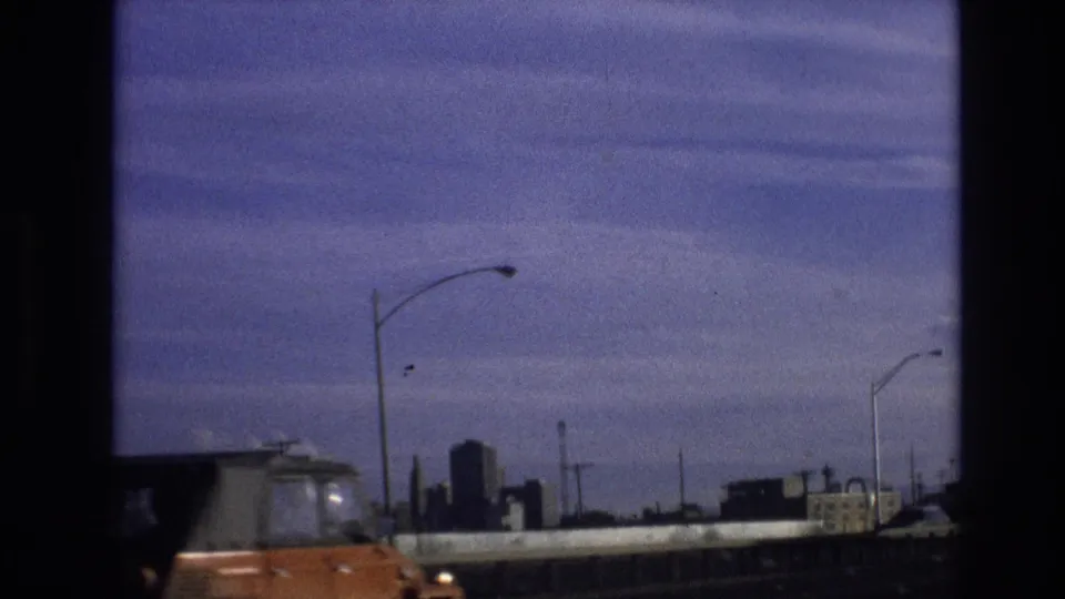 Final 1974 8mm frame from the supplied Vail, Colorado clip showing an orange vehicle near the camera, streetlights, highway barriers, and a distant skyline.