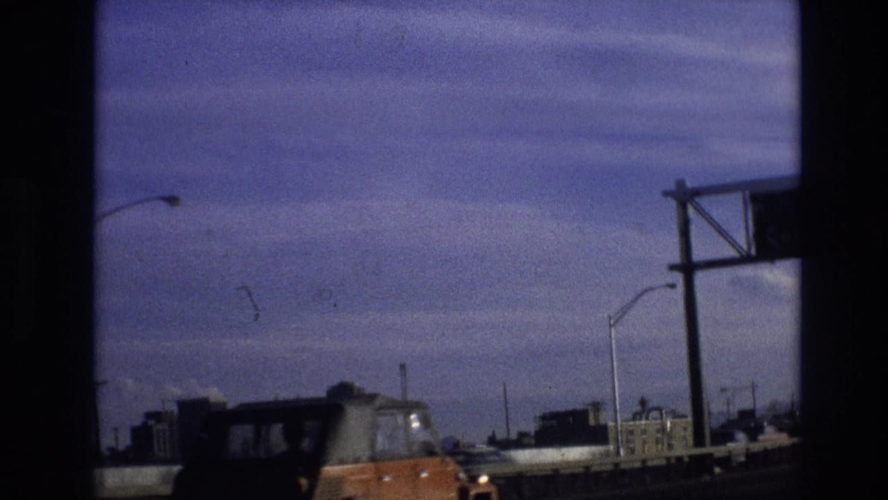 Orange car and other vehicles pass beneath a dark bridge frame with distant buildings and streetlights.