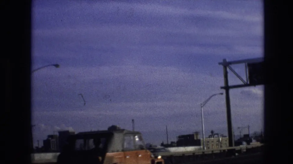 1974 8mm frame from Vail, Colorado showing traffic on a bridge or highway, with an orange car, streetlights, distant buildings, and heavy shadow from the filming position.