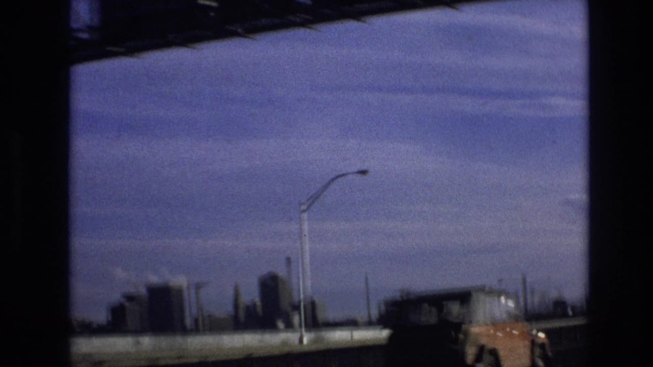 1974 8mm roadway view in Vail, Colorado with a tall streetlight, low distant skyline, guardrail, blue-tinted sky, and the partial front of an orange vehicle at frame edge.