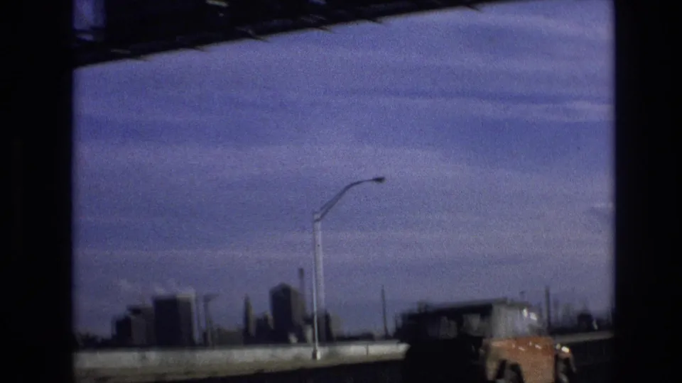 1974 8mm roadway view in Vail, Colorado with a tall streetlight, low distant skyline, guardrail, blue-tinted sky, and the partial front of an orange vehicle at frame edge.