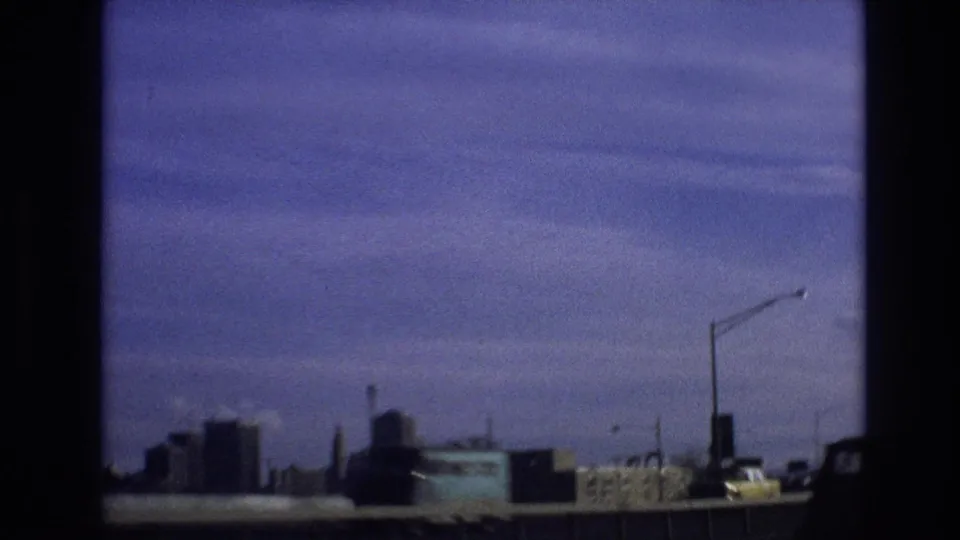 1974 Vail, Colorado home-movie frame showing a moving-vehicle perspective across a bridge or highway, with streetlight poles, passing vehicles, guardrail, and distant buildings.