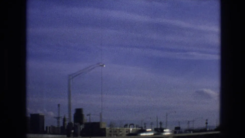 1974 8mm frame from a moving vehicle in Vail, Colorado showing bridge or highway infrastructure, tall streetlights, a guardrail, distant structures, and blue sky.