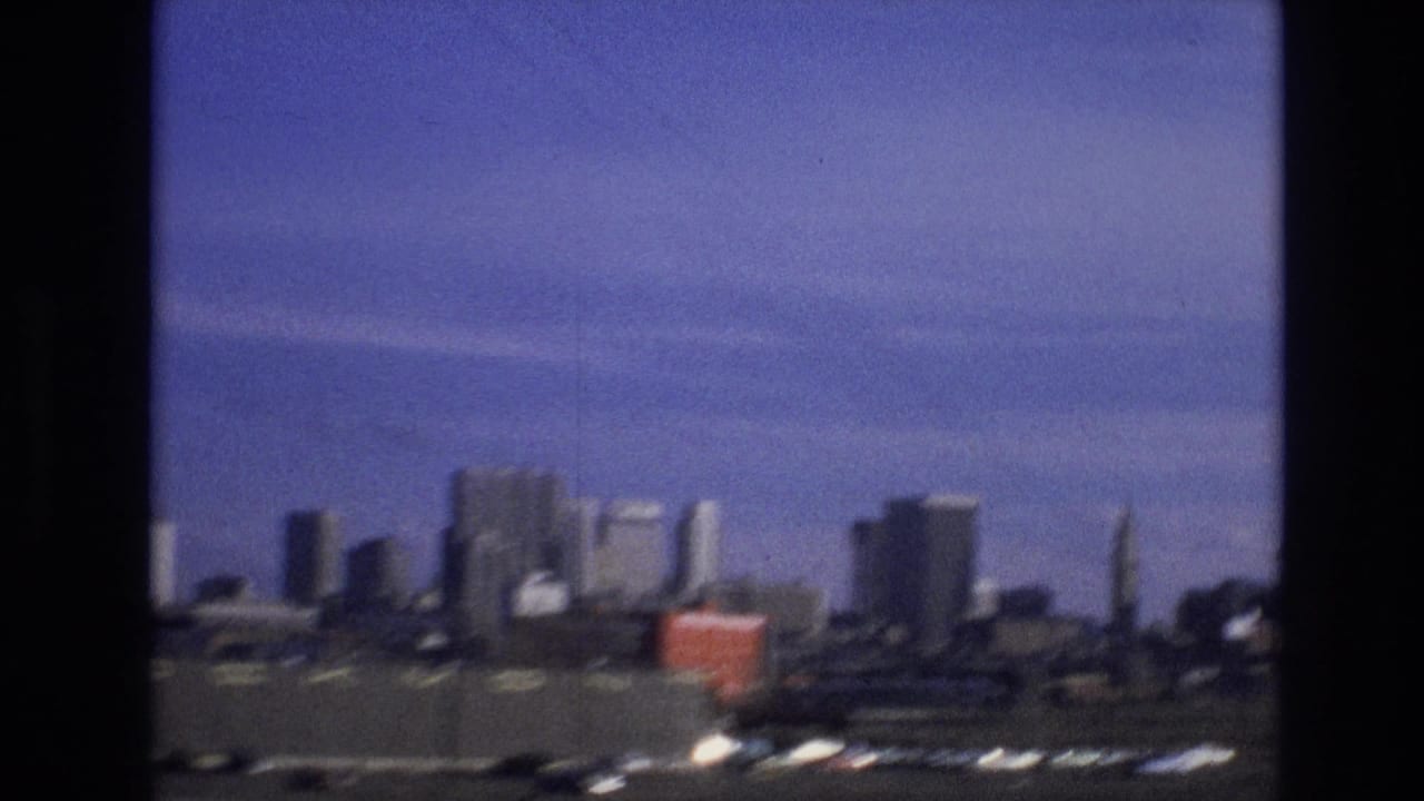 Soft 8mm frame of a distant city skyline with industrial foreground, a red container-like object, and dark window or film borders from a moving travel shot.