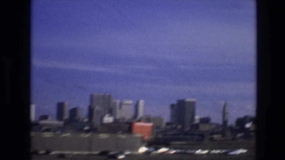 Soft 8mm frame of a distant city skyline with industrial foreground, a red container-like object, and dark window or film borders from a moving travel shot.