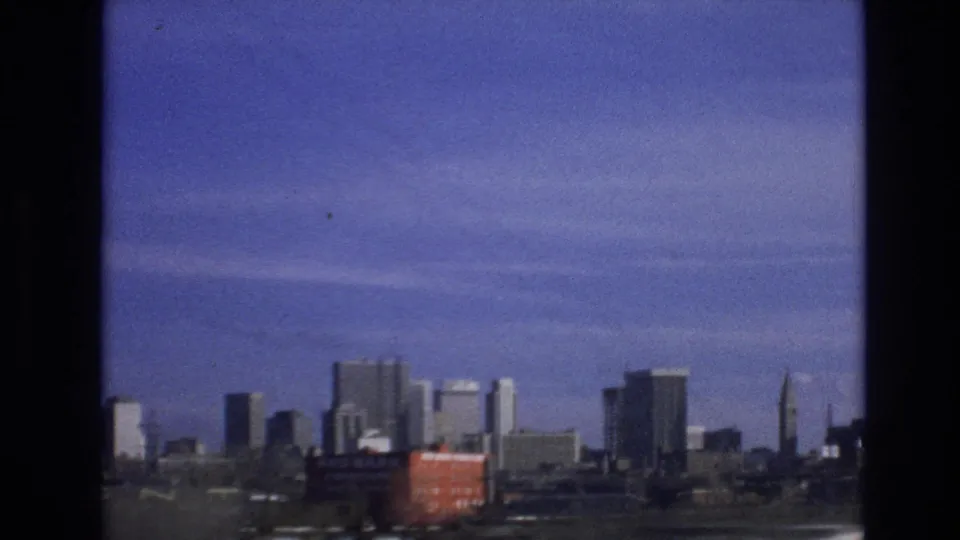 1974 home-movie frame showing a broad skyline under blue sky, with a red container-like object and low industrial area in the foreground.