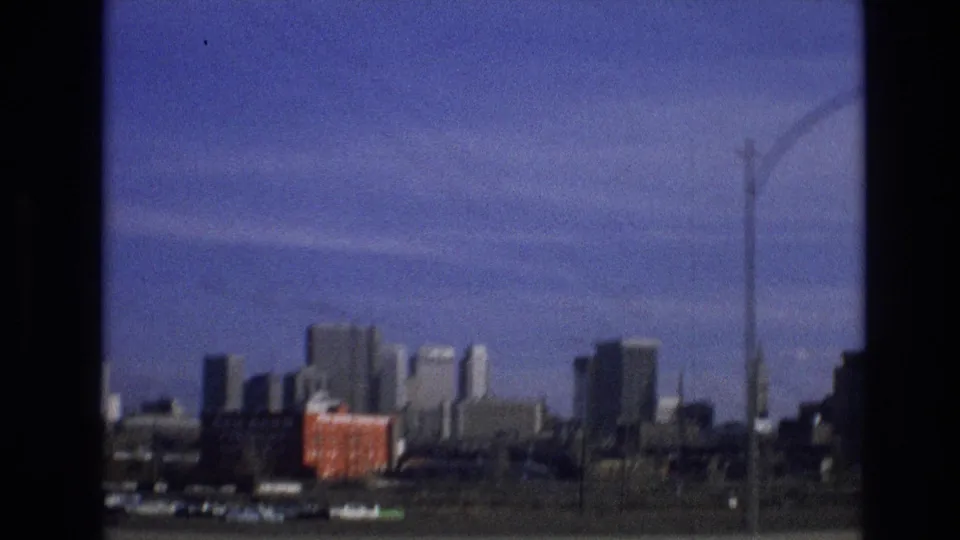 1974 8mm frame showing a distant urban skyline, a crane at the right edge, and a red container-like object in the industrial foreground.