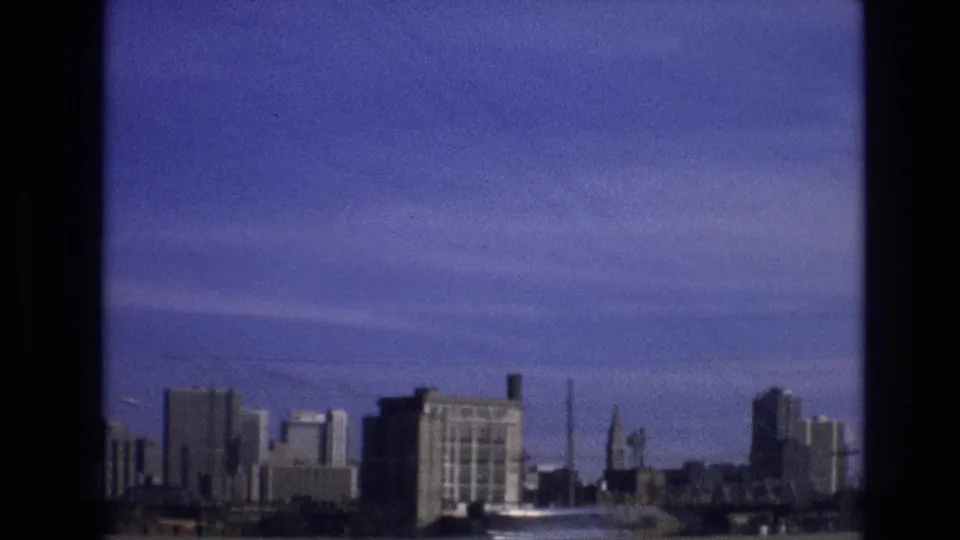 1974 Colorado 8mm frame showing a distant city skyline, tall buildings, a roadside pole, and blue sky from a moving travel viewpoint.