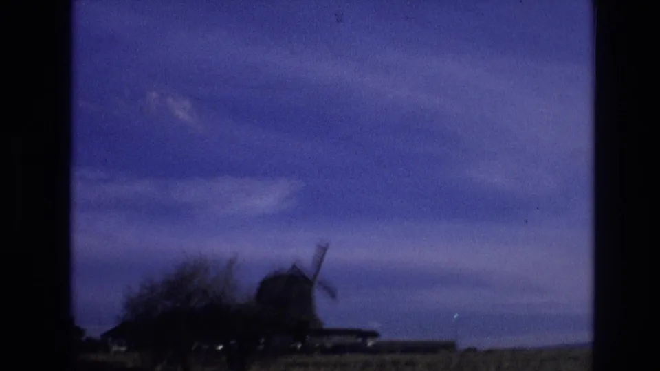 1974 8mm Vail, Colorado travel frame showing a roadside view of a windmill, low rural buildings, open field, and dark foreground edges under a blue evening sky.