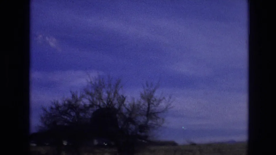 1974 8mm Vail, Colorado frame with leafless trees, low countryside structures, and a dark foreground silhouette passing across a blue sky during a roadside drive.