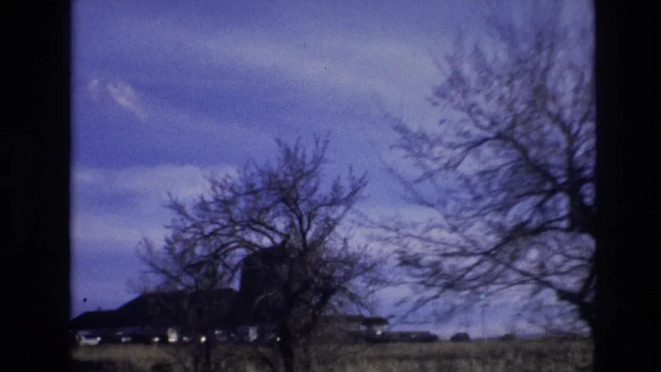 1974 8mm Vail, Colorado travel frame showing leafless roadside trees, an open field, low farm-like buildings, and soft vehicle-motion blur beneath a blue sky.