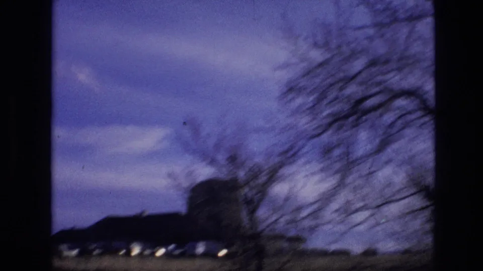 1974 8mm Vail, Colorado frame with a blurred leafless tree in the foreground, a dark low building silhouette, open field, and blue twilight sky from a moving vehicle.