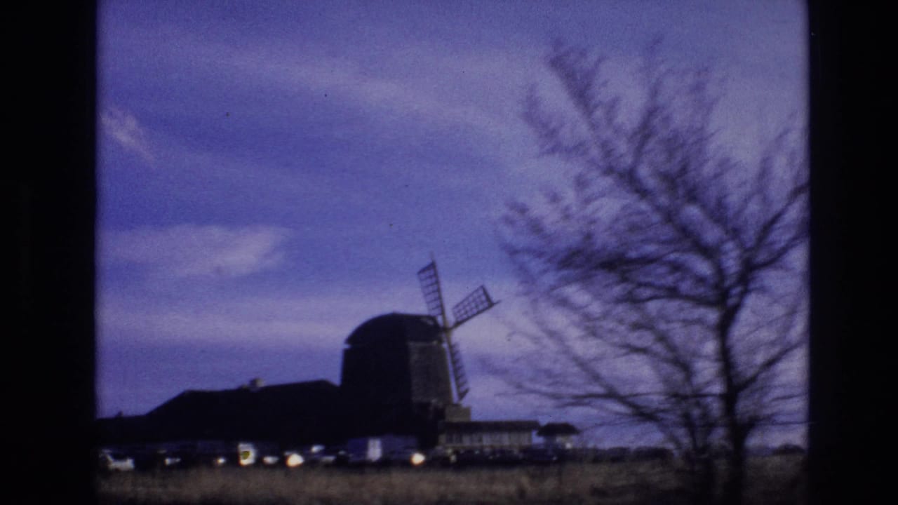 1974 Vail, Colorado home-movie frame showing a silhouetted windmill and low buildings beyond a field, partly framed by bare tree branches under a purple sky.