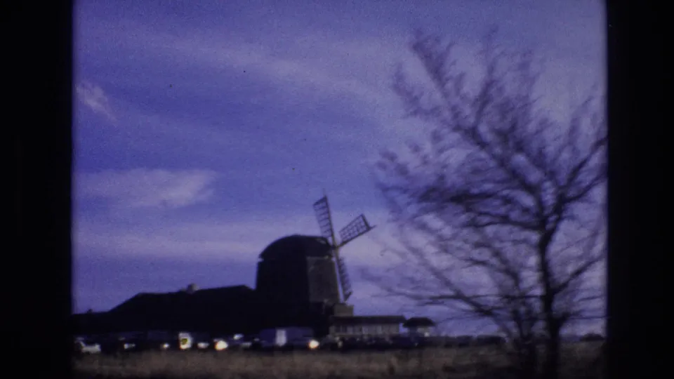 1974 Vail, Colorado home-movie frame showing a silhouetted windmill and low buildings beyond a field, partly framed by bare tree branches under a purple sky.