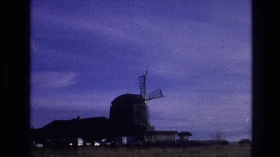 1974 Vail, Colorado frame with a dark windmill silhouette, adjacent low buildings, faint lights near the ground, and open sky viewed from a moving vehicle.