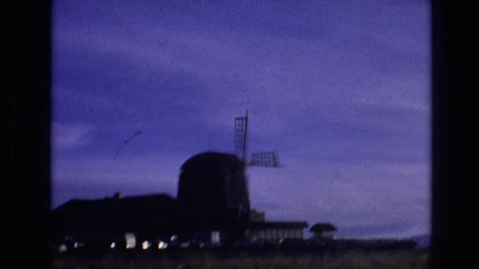 1974 Vail, Colorado home-movie frame showing the windmill partly blurred by motion, with dark rooflines, field grass, small lights, and a broad purple sky.