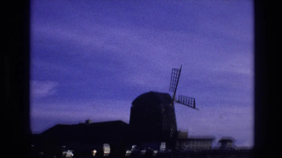 1974 Vail, Colorado frame showing a large windmill beside a low dark building, with field grass, faint lights, and blue-purple evening sky in a grainy 8mm scan.