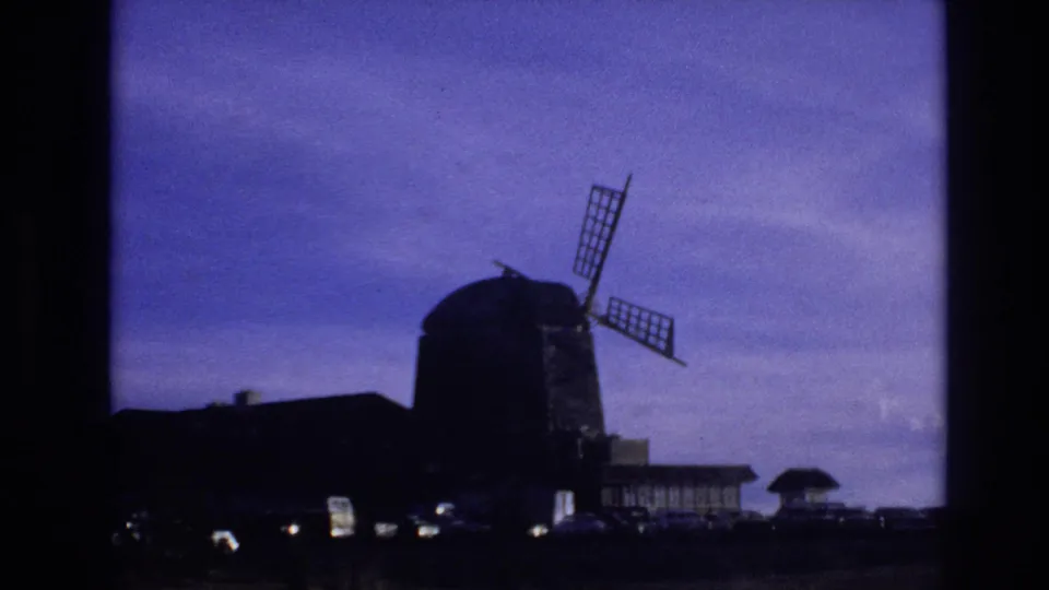 1974 Vail, Colorado home-movie frame showing a large silhouetted windmill near parked cars and low buildings under a blue sky.