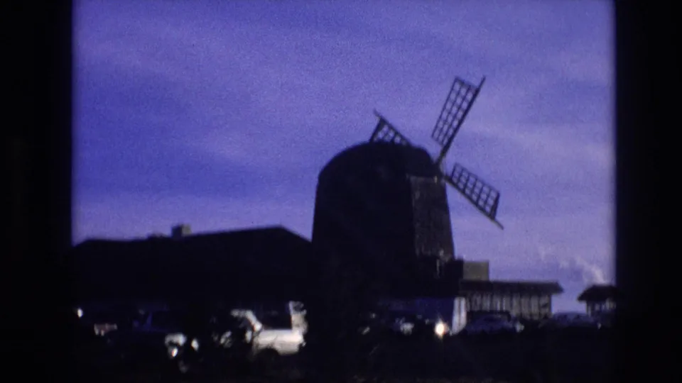 A traditional windmill in Vail, Colorado appears in silhouette as the camera passes a parking lot filled with period cars.