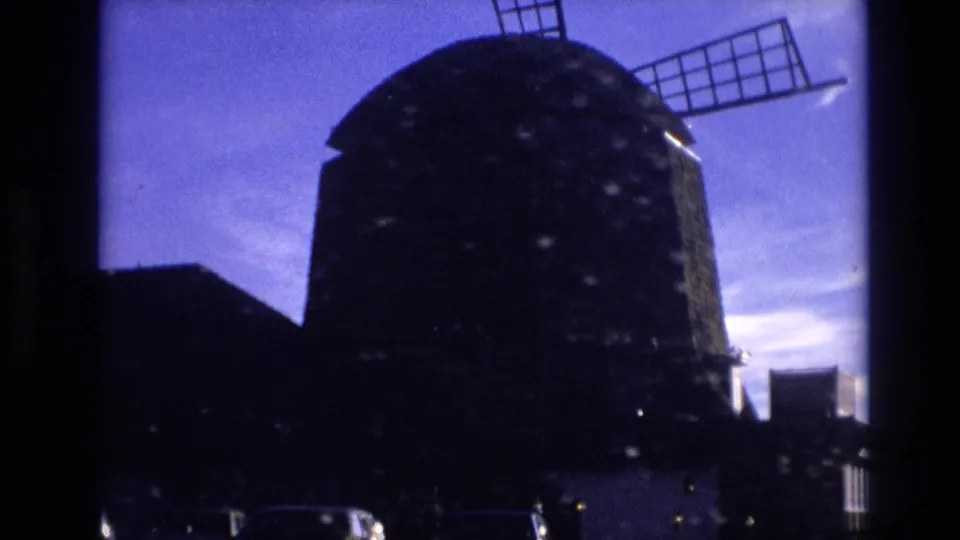 Closer 8mm frame of the dark windmill tower and blade structure in Vail, with a car and building edges visible below.
